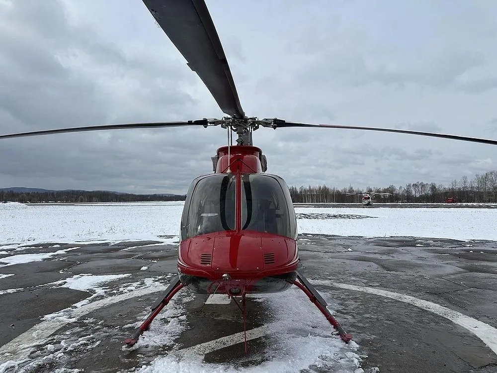 A front view of a red Bell 407GXP helicopter parked on a snowy helipad, with another helicopter in the distant background.