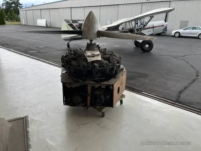 A large radial aircraft engine with a propeller is on a wooden crate outside a hangar, with a white and black small aircraft in the background.