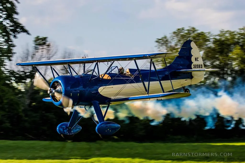 A blue and white biplane with registration N32063 flying low over a grassy field, emitting smoke.