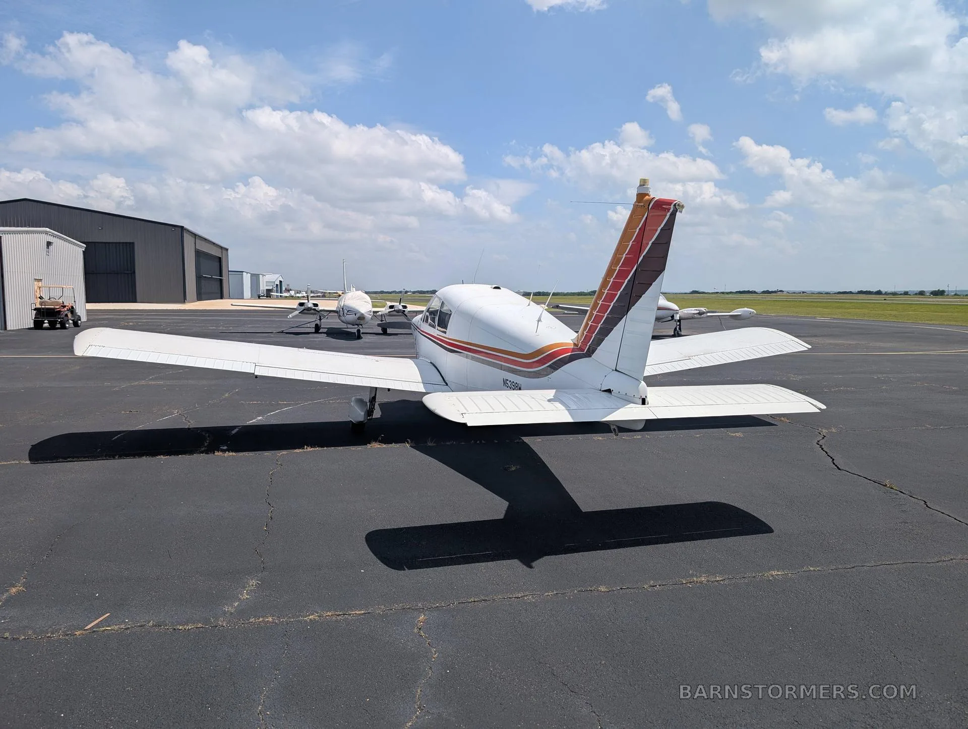 A white single-engine low-wing aircraft with red, orange, and brown stripes on the tail and fuselage, viewed from the rear, parked on an asphalt ramp.