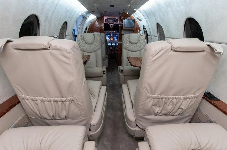 The interior cabin of a private jet, showing beige leather seats and a view down the aisle towards the cockpit.