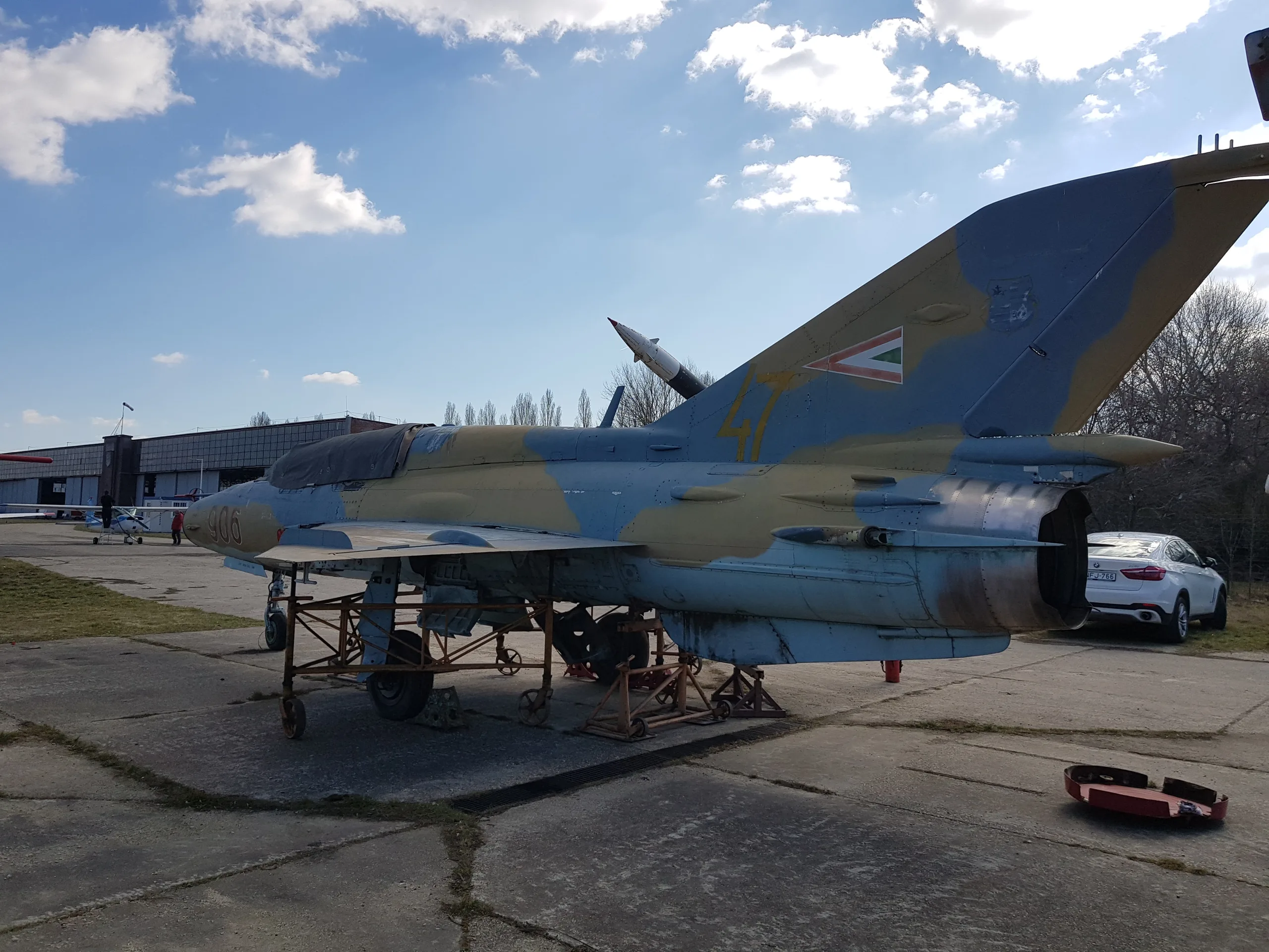 A side view of a MiG-21 fighter jet in blue and tan camouflage with Hungarian markings on an airfield, with a building in the background.
