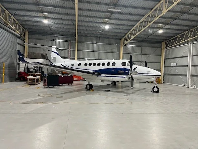 A white and blue twin-engine turboprop aircraft, likely a Beechcraft King Air, is parked inside a hangar.