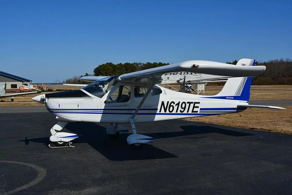 A white Tecnam Light Sport aircraft with blue and grey stripes parked on an asphalt ramp.