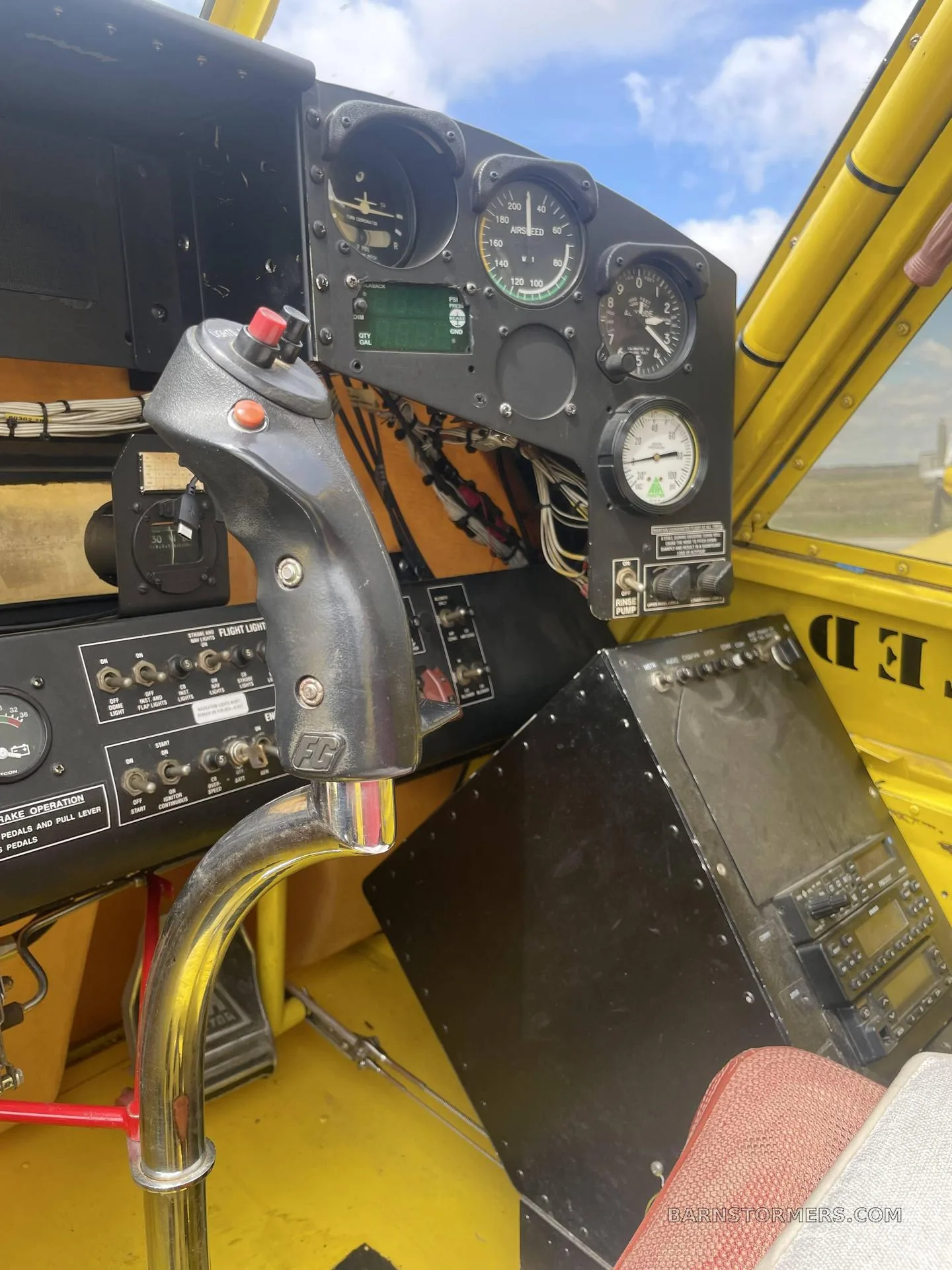 The cockpit interior of a yellow agricultural aircraft, showing the control stick, instrument panel, and avionics stack.