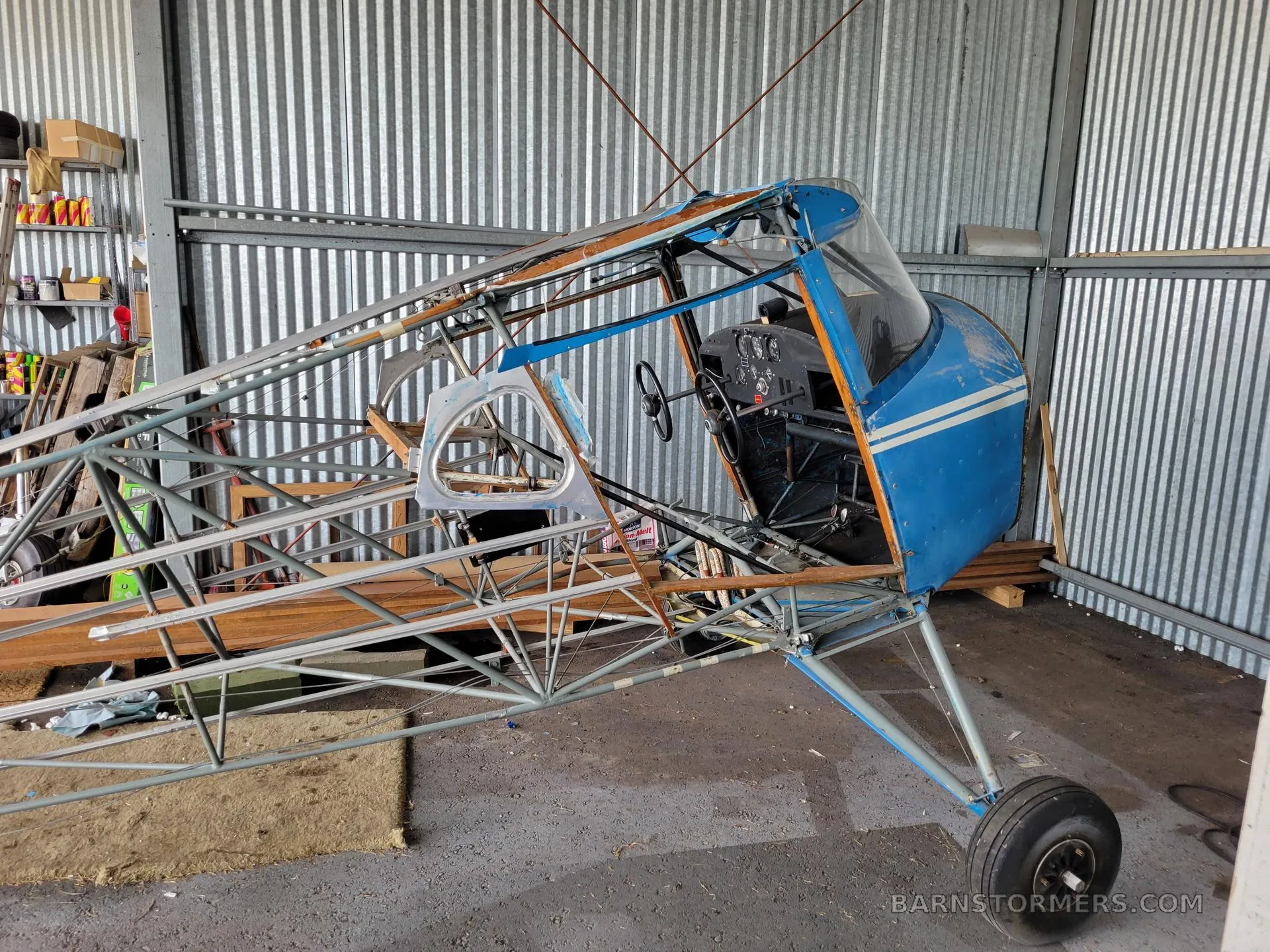A partially assembled blue and white aircraft fuselage and frame in a corrugated metal hangar.