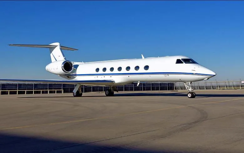 A white Gulfstream business jet with a blue stripe on its fuselage is parked on an airport tarmac under a clear blue sky.