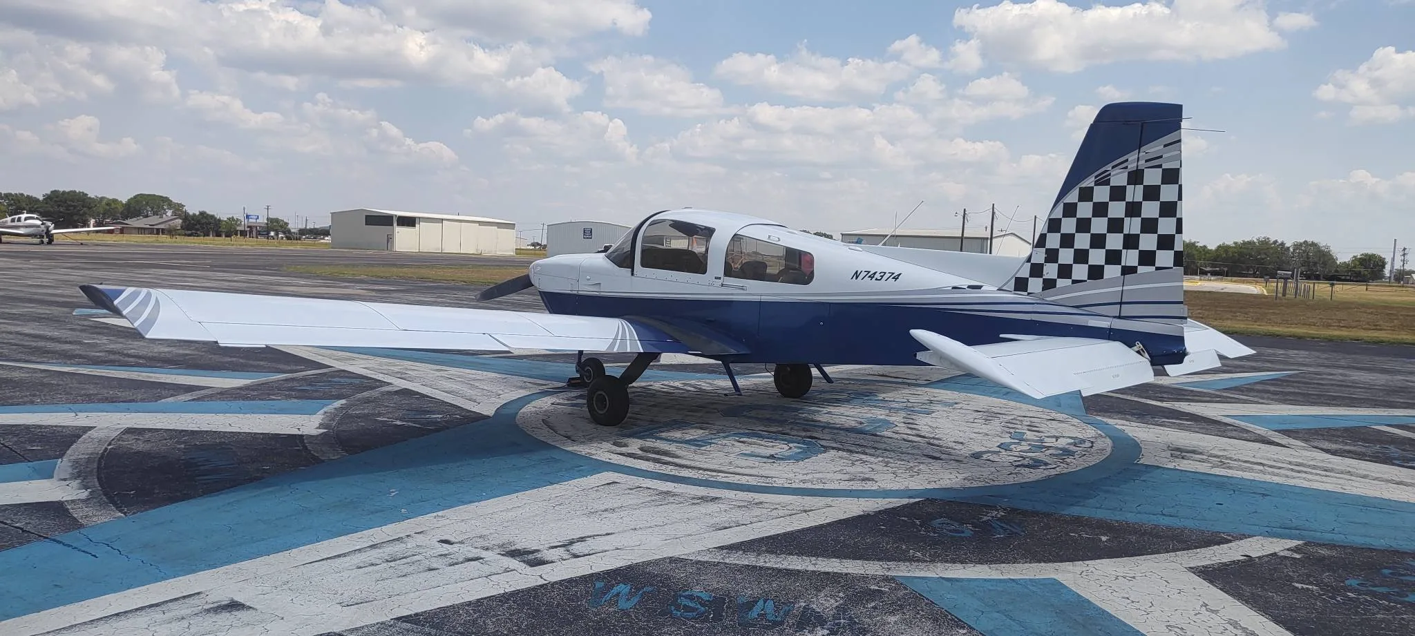 A blue and white single-engine aircraft, N74374, with a checkered tail, parked on an airfield compass rose under a partly cloudy sky.