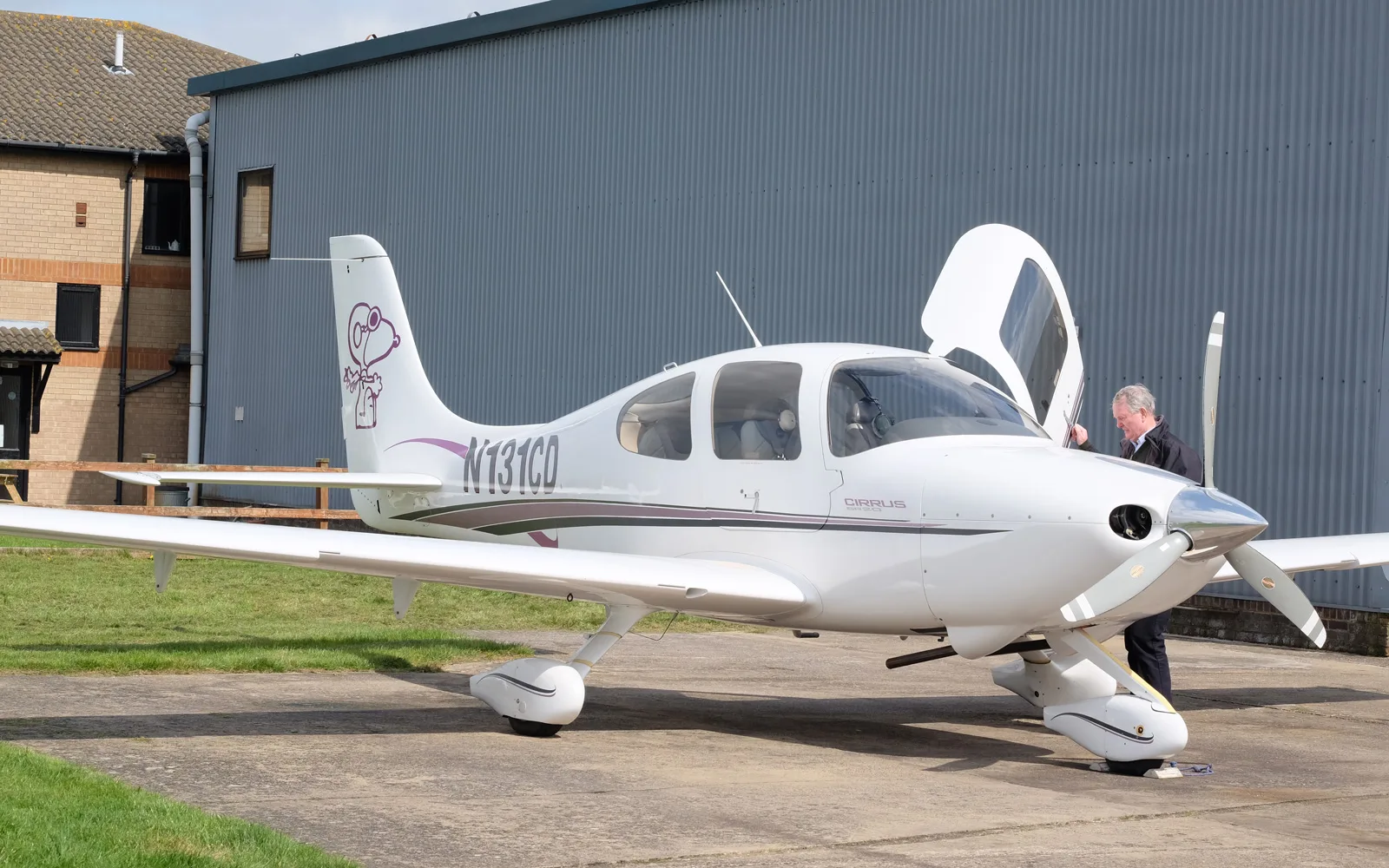 A white Cirrus SR22 aircraft with open door and a Snoopy graphic on the tail, with a person standing near the propeller.