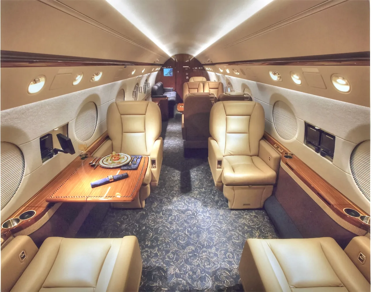 Aircraft cabin interior with leather seats, wooden table, and blue carpet.