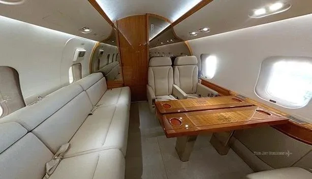 A view inside a private jet cabin showing a long beige sofa on the left and two individual seats with a wooden table on the right.