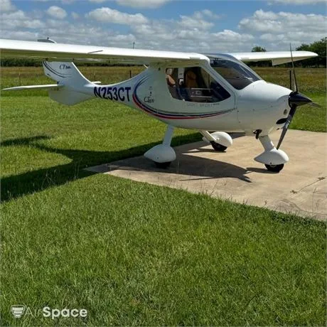 A white Flight Design CT light-sport aircraft with red, white, and blue stripes, registration N253CT, parked on a concrete pad next to a grassy field.