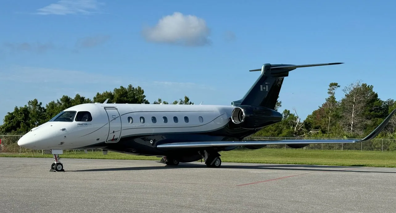 A white and dark grey Embraer Praetor 600 private jet with a Canadian flag on its tail is parked on a tarmac under a blue sky.