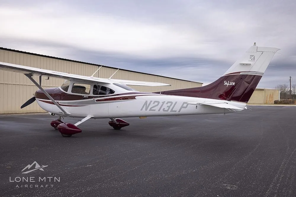 A Cessna 182 Skylane with a maroon and white paint scheme, parked on an asphalt ramp next to a hangar under a cloudy sky, viewed from the rear-left.