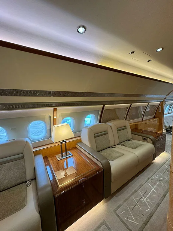 A section of a private jet cabin features two-seater sofas, a polished wooden table with a lamp, and large windows.