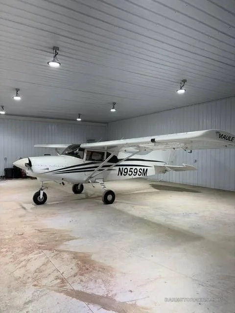 A white Maule M-7-235B aircraft with black stripes and registration N959SM is parked inside a hangar, viewed from the front-left.