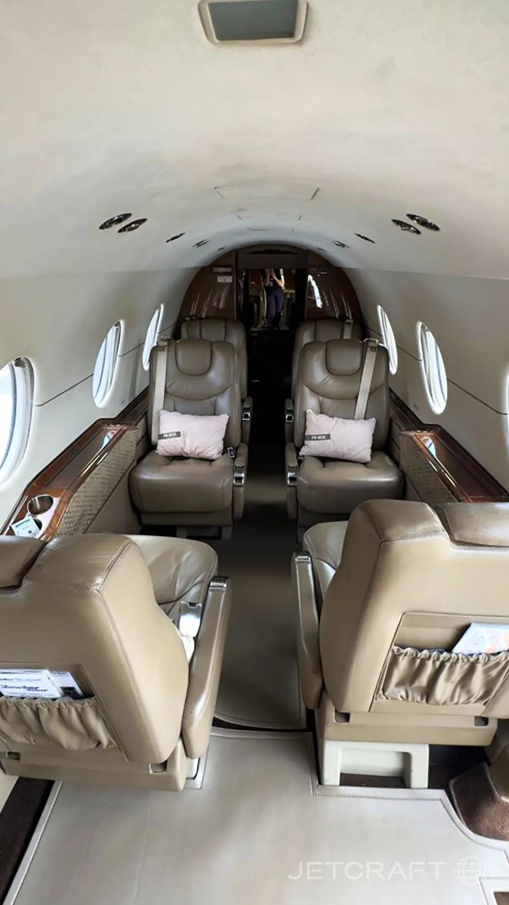 The interior of a private jet cabin features tan leather seats arranged in a club configuration with small pillows.