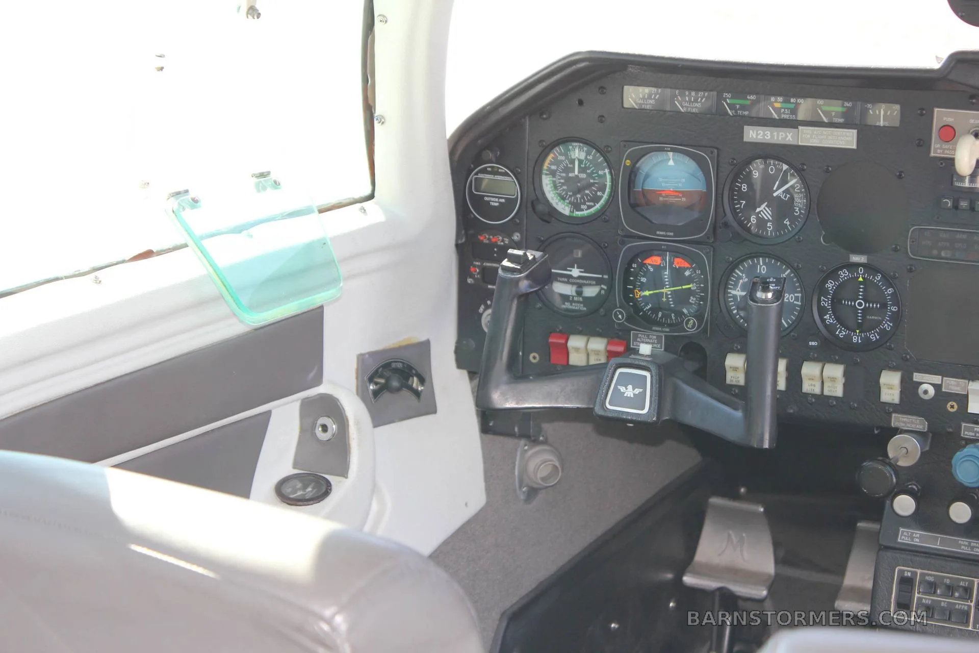 The cockpit of a Mooney M20J, showing the instrument panel with analog gauges, a black yoke, and a side window.