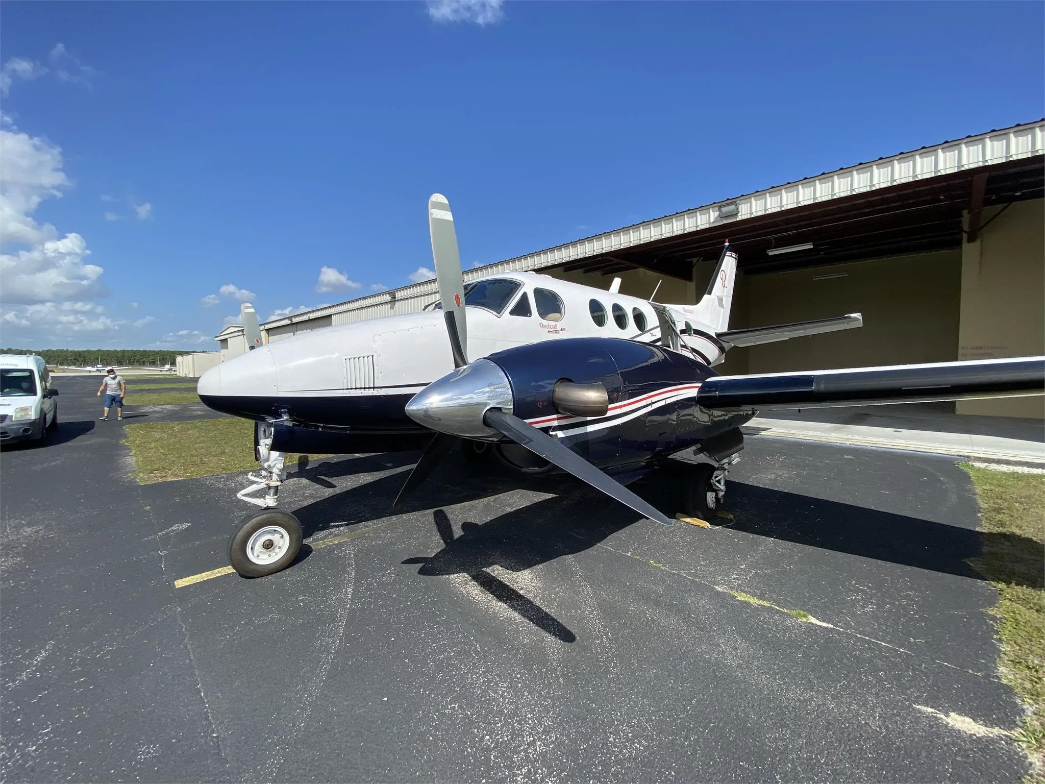 A front-quarter view of a white and dark blue Beechcraft King Air with propellers, parked on an asphalt apron with a person in the background.