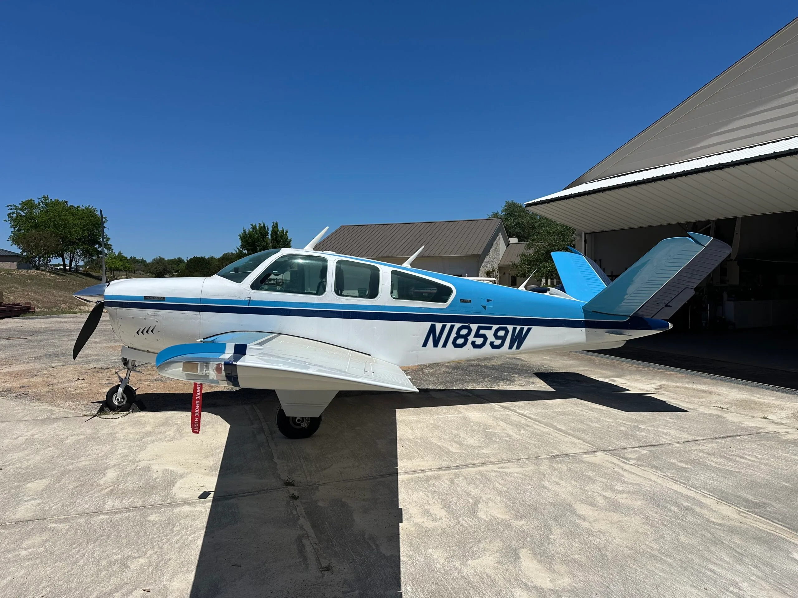 A white and blue Beechcraft Bonanza with registration N1859W is parked on a concrete pad next to an open hangar under a clear blue sky.