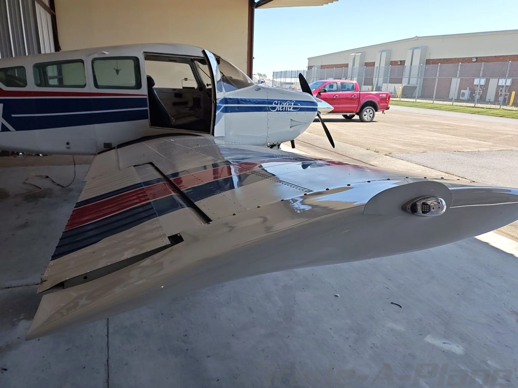 A white Beechcraft Sierra N23PX with blue and red stripes parked in a hangar with the pilot's door open, viewed from the front right wing.