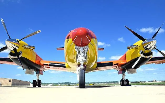 A head-on view of the King Air 350 aircraft, showing its distinctive orange, yellow, and blue 'Just for Kids' paint scheme and twin propellers.