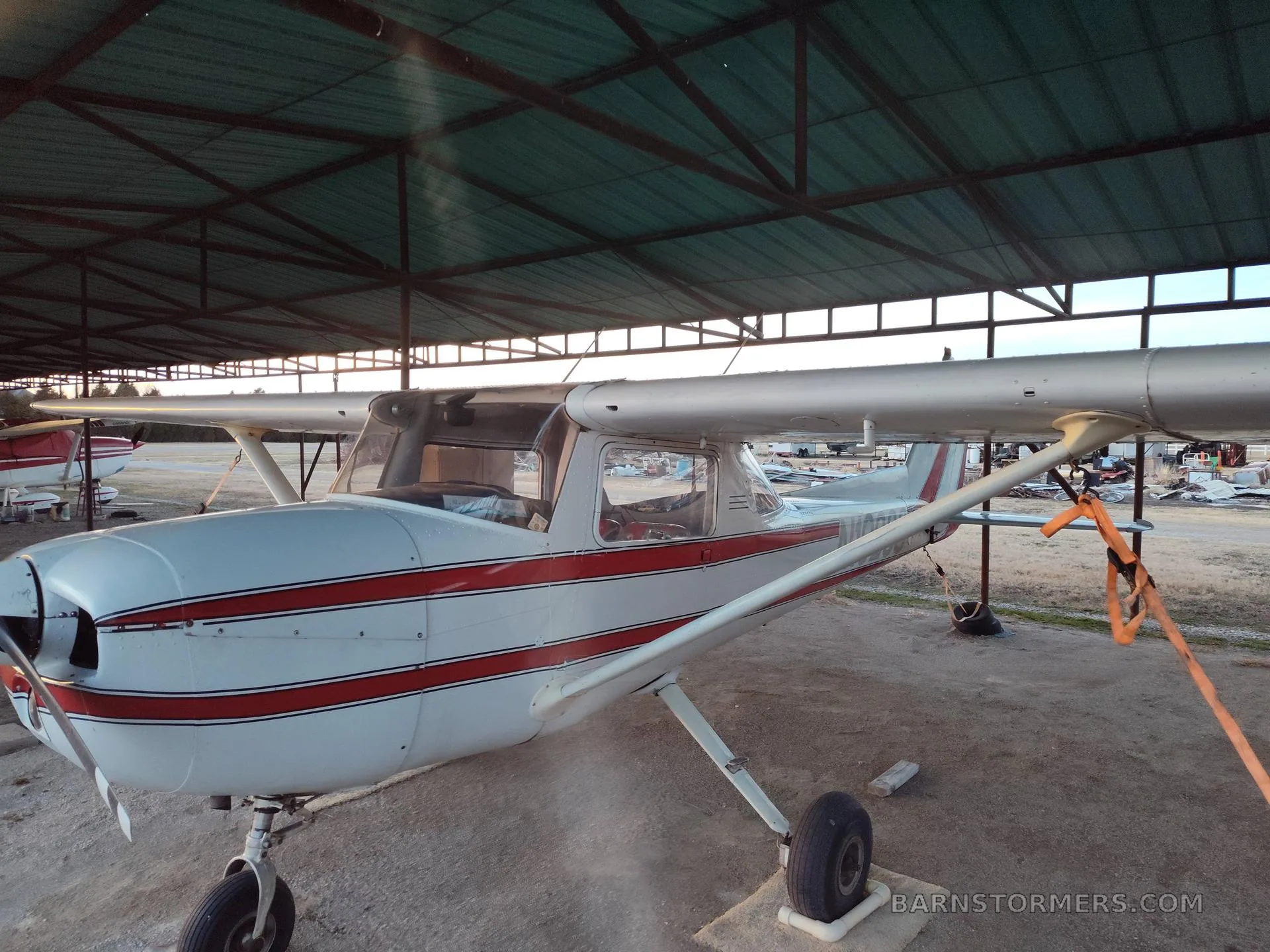 A white and red striped Cessna 150/152 type aircraft is parked under a metal hangar roof.