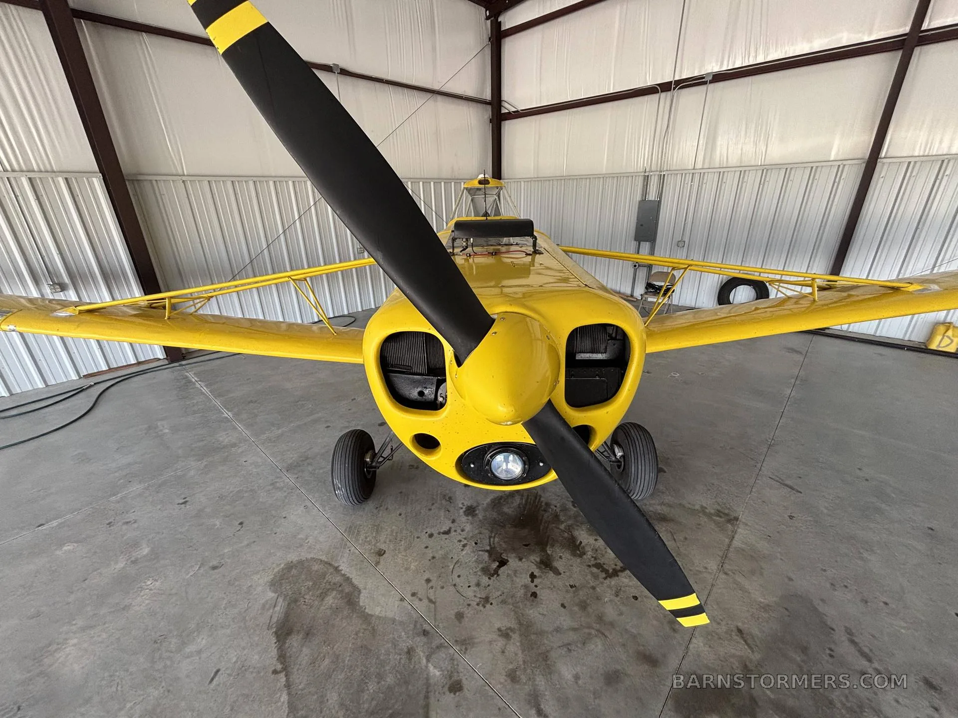 Front view of a yellow Piper PA-25 Pawnee aircraft with a black propeller, inside a hangar.