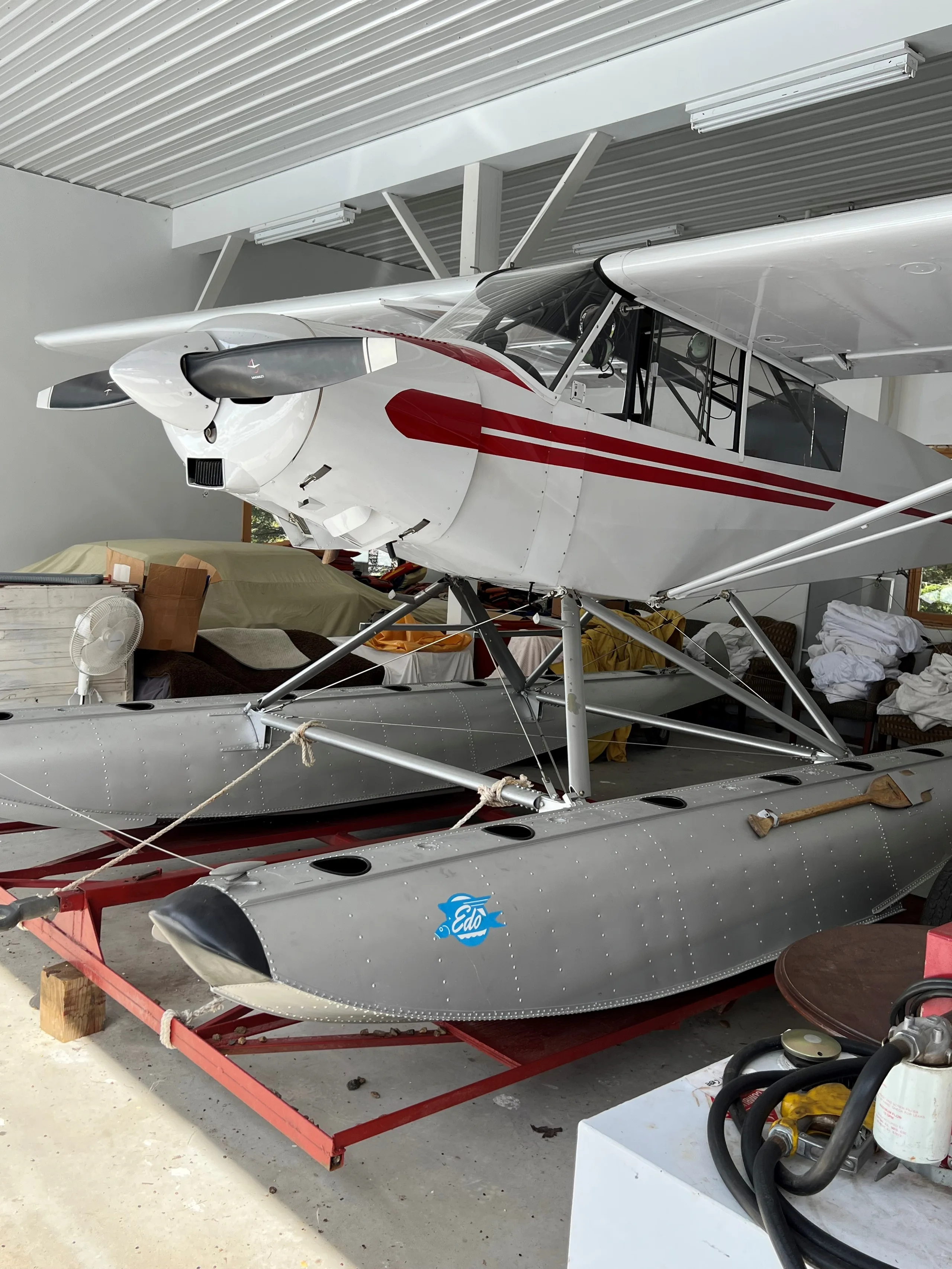 A white floatplane with red stripes is parked indoors on a red dolly, viewed from the front-right, showing its Edo floats and a paddle.