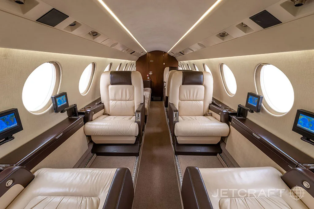 The luxurious cabin interior of a business jet with light beige leather seats and individual entertainment screens.