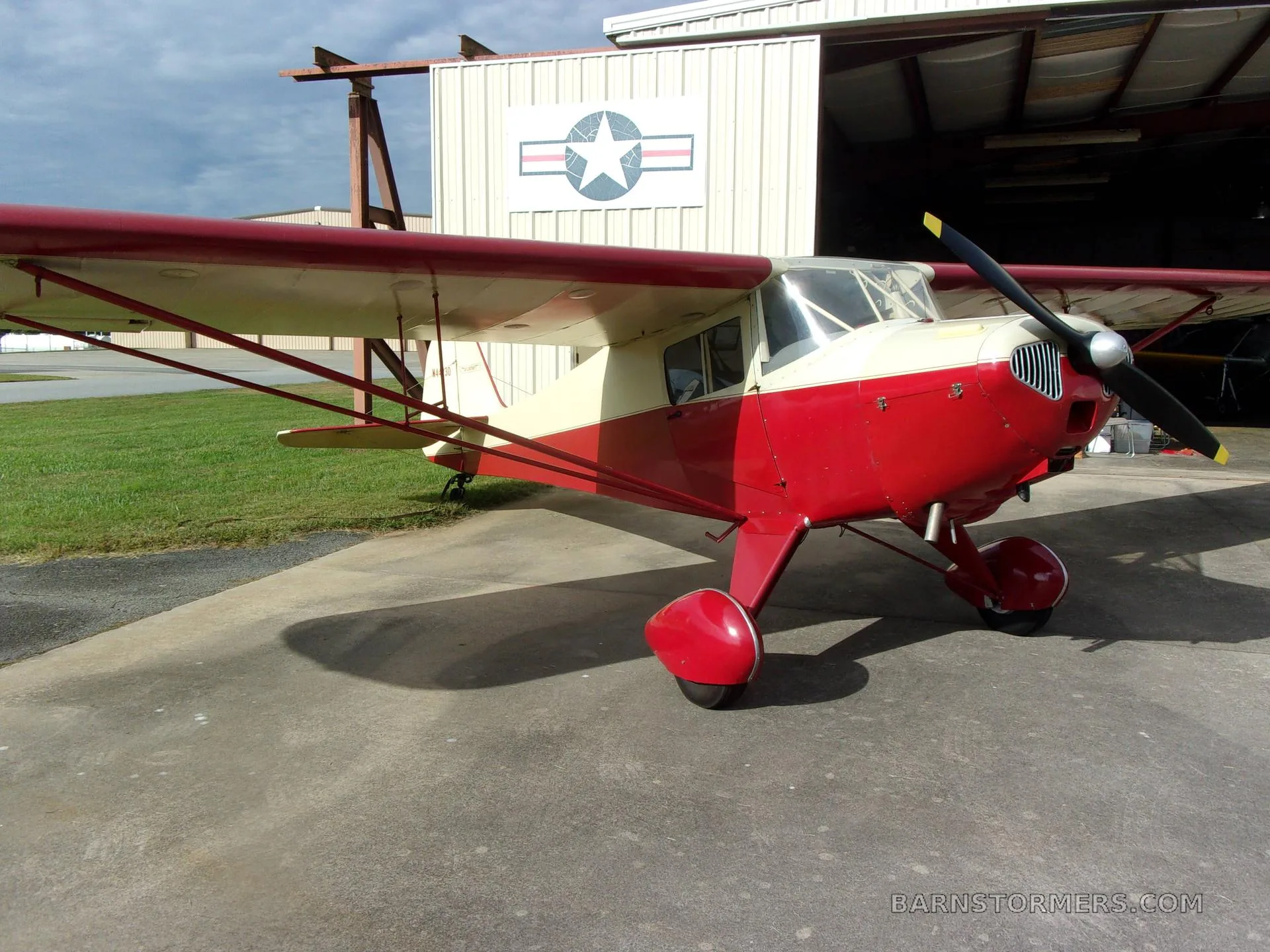 A red and cream Taylorcraft BC-12D aircraft parked on a concrete apron outside a hangar, viewed from the front-right.