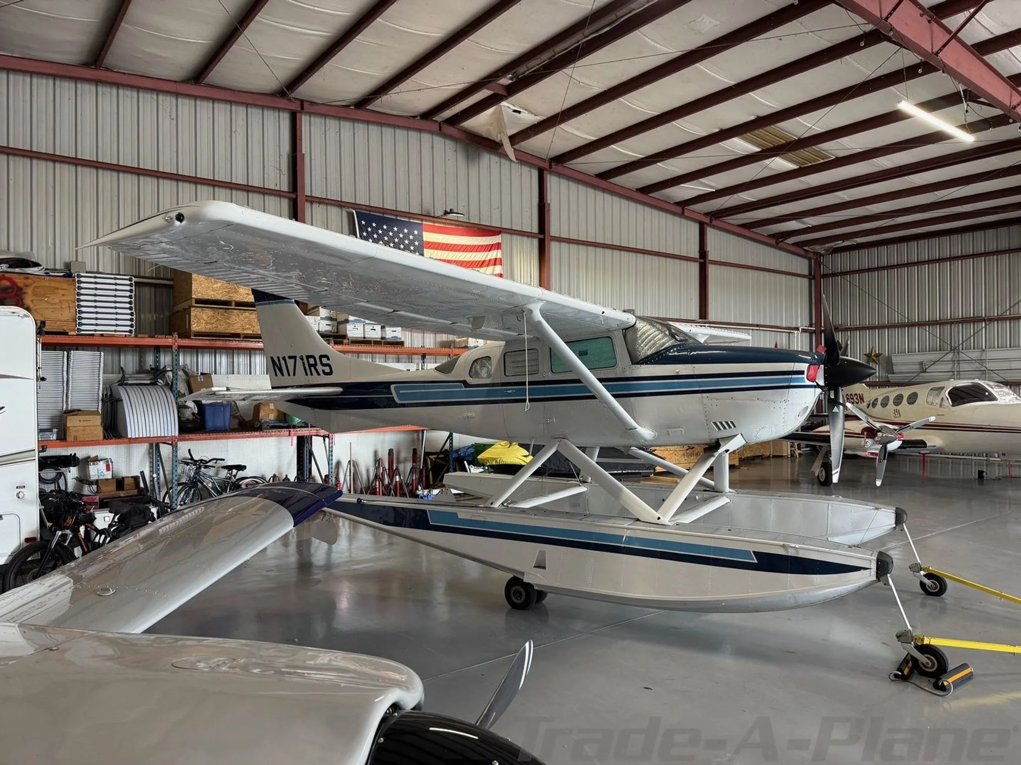 A white and blue Cessna 206 Stationair floatplane, registration N171RS, parked in a hangar.