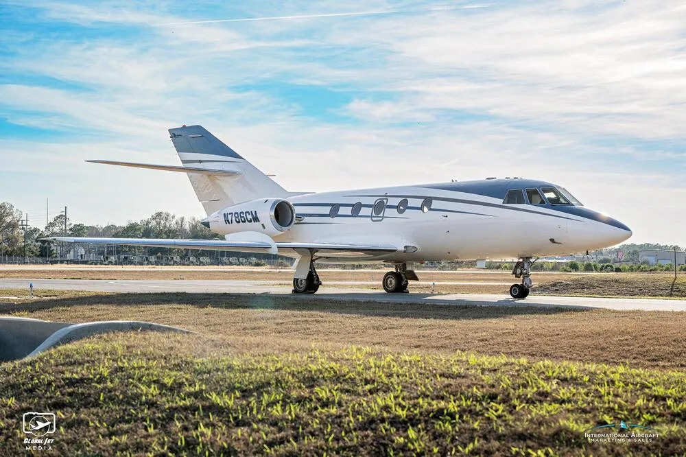A side profile of the white and blue business jet N786CM on a tarmac with a grassy foreground.