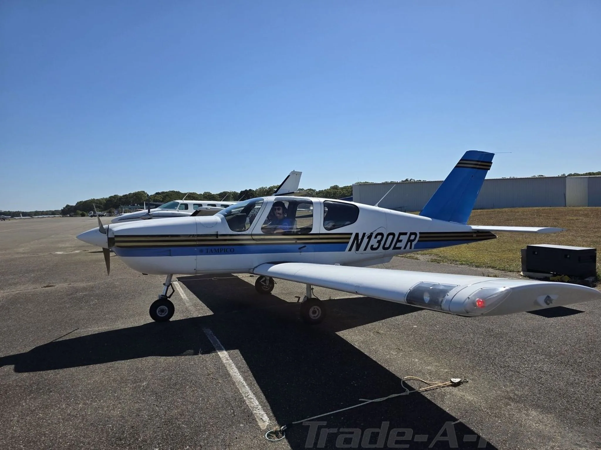A white and blue Socata TB-9 Tampico aircraft with tail number N130ER parked on an asphalt ramp on a sunny day.