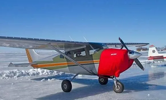 A Cessna 182D aircraft with a green, white, and orange stripe paint scheme, a red engine cover, parked on a snowy or icy surface.