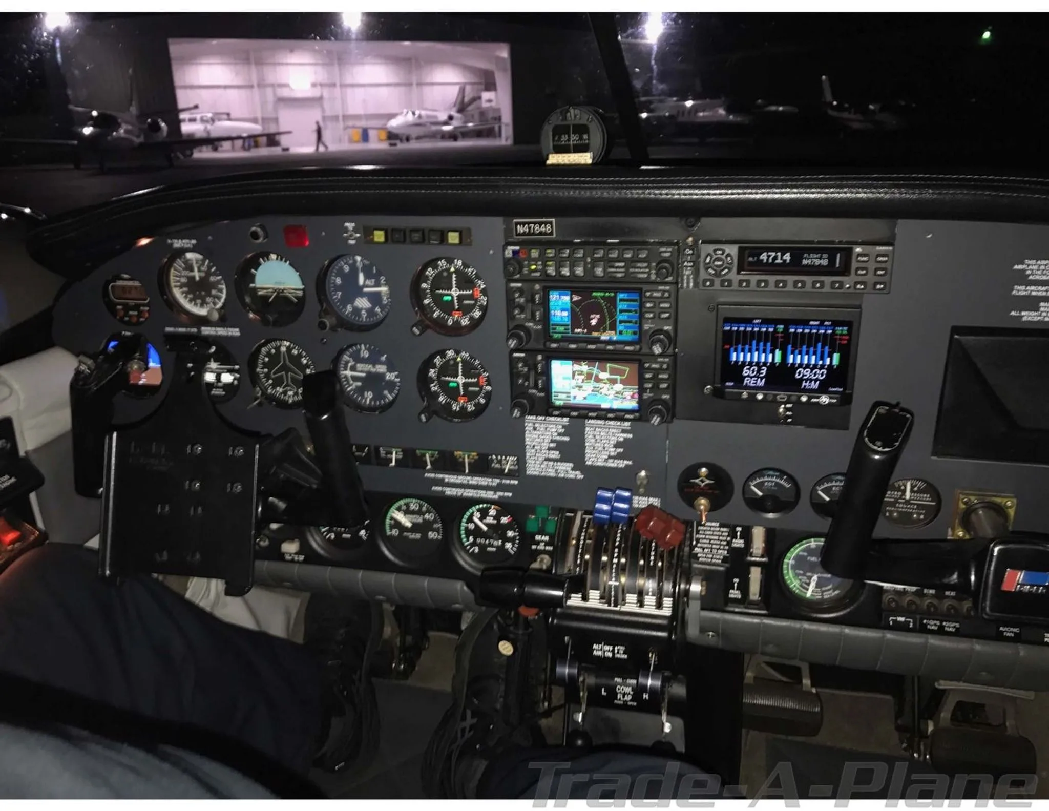 A view of the aircraft's cockpit at night, showing the instrument panel with multiple gauges and digital displays, with other aircraft visible in a hangar in the background.