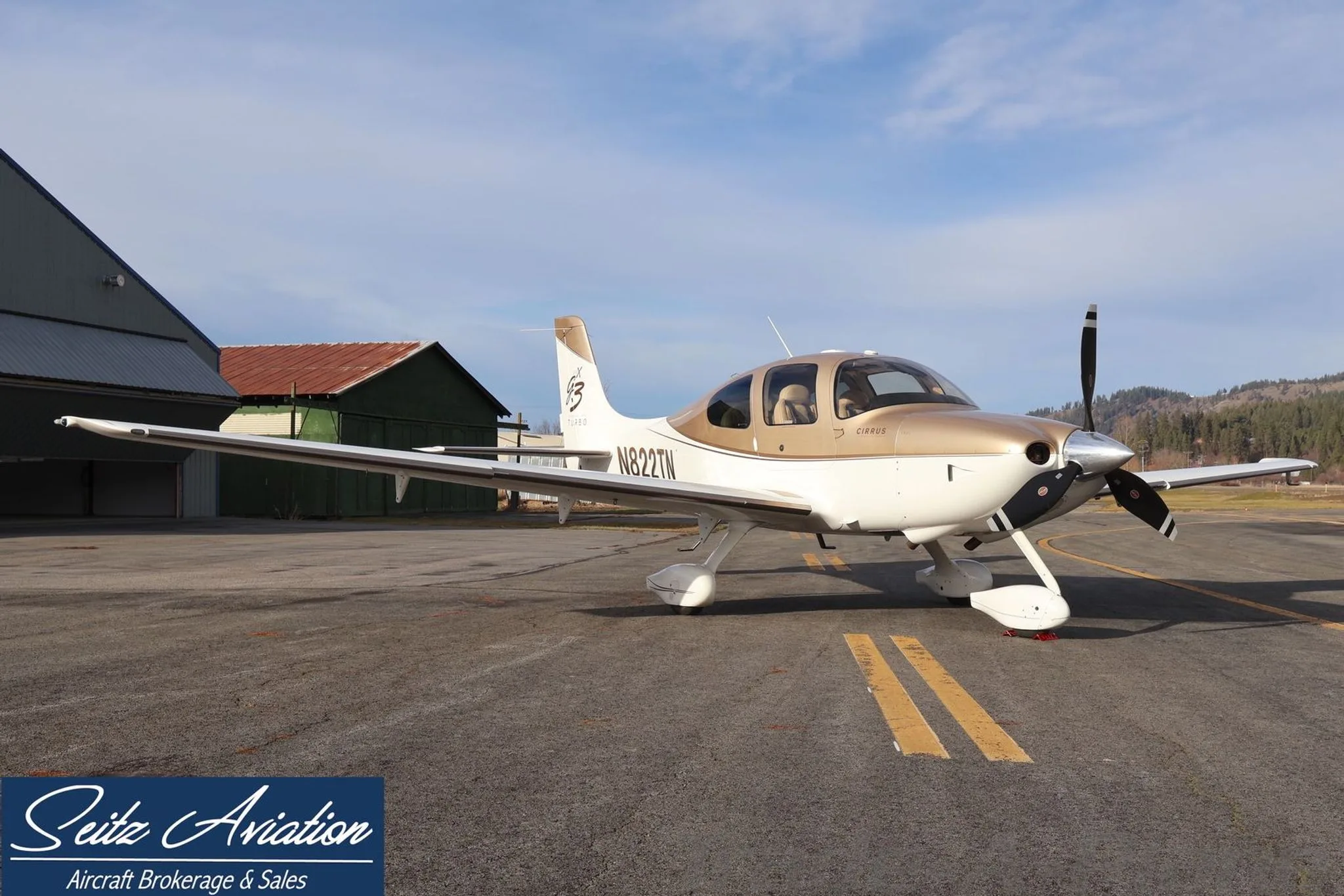 A Cirrus SR22T aircraft with N822TN registration, painted white and gold, parked on an asphalt ramp with hangars in the background.
