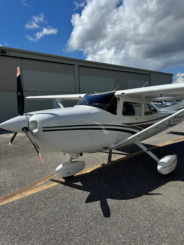 Side-front view of a white Cessna 182 Skylane with black and brown stripes, parked on an asphalt ramp next to a hangar.