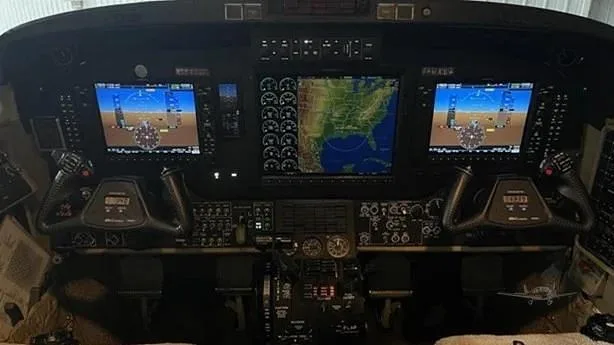 A detailed view of an aircraft cockpit with multiple digital displays showing navigation and flight information, and two control yokes.