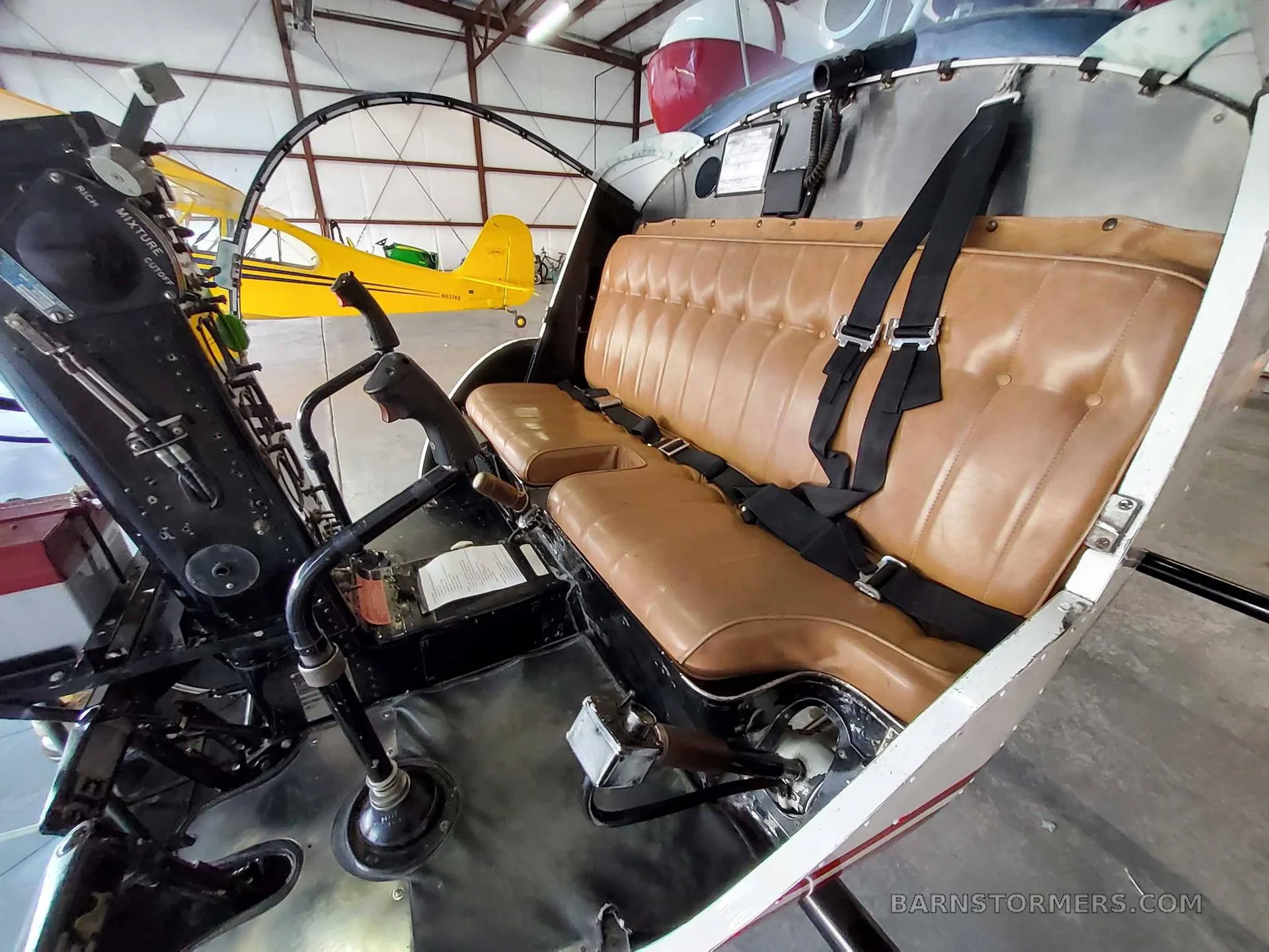 The tan leather bench seat and controls inside a helicopter cabin, with another yellow aircraft visible in the hangar background.