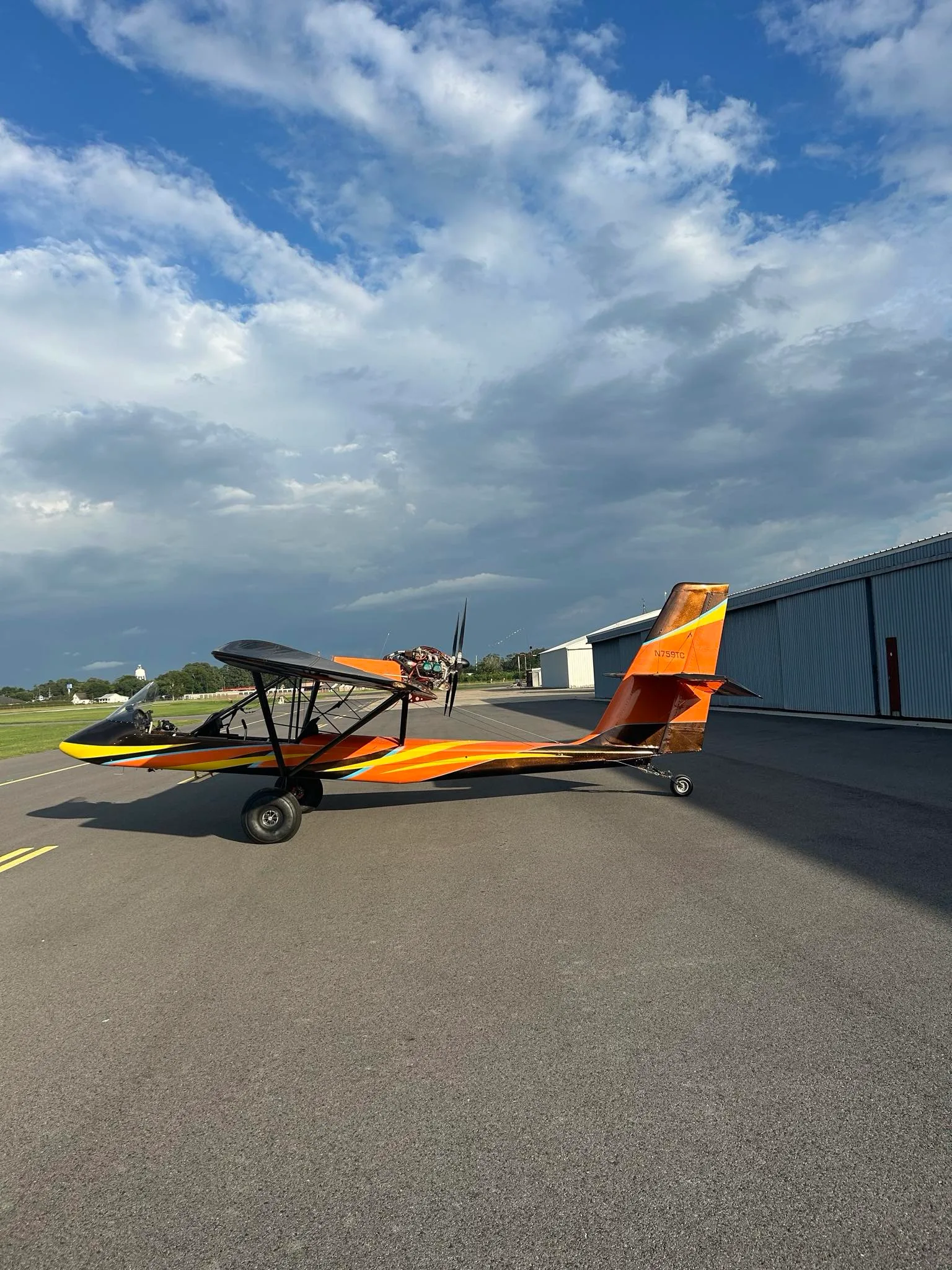 A side view of an orange and black ultralight aircraft with a unique paint scheme, parked on an asphalt runway with hangars in the background under a cloudy sky.