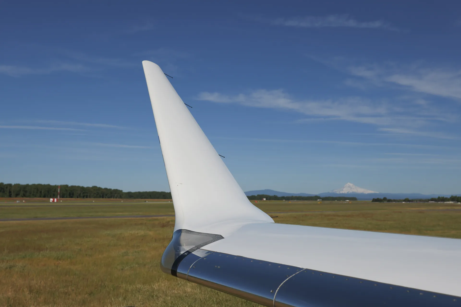A close-up of the white wingtip and polished leading edge of a business jet's wing, with a distant mountain under a blue sky.