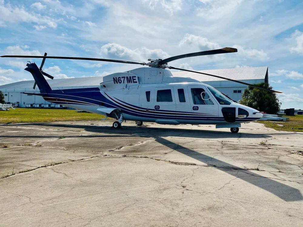 Side view of a white and blue helicopter (N67ME) with red stripes parked on a concrete pad next to a hangar.