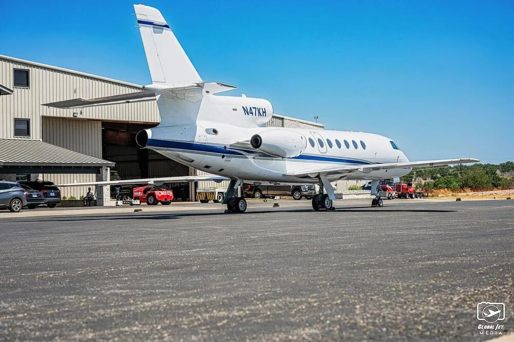 A rear-side view of a white Dassault Falcon 900EX business jet with blue stripes, parked on an airfield next to a hangar.