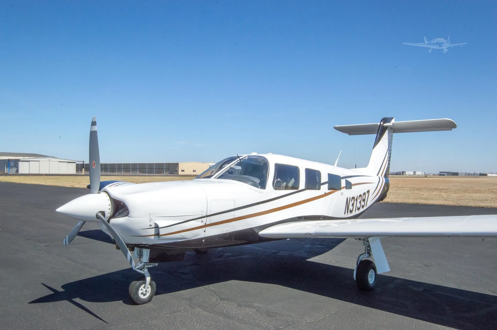 A white single-engine propeller aircraft with brown and black stripes, registration N31397, is parked on a tarmac with hangars in the background under a clear blue sky.