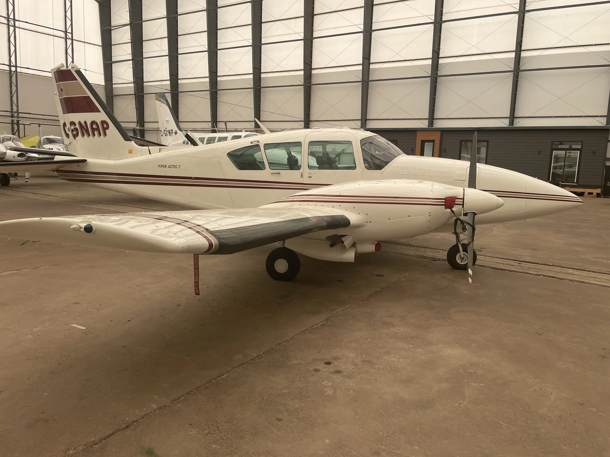 A white Piper Aztec F (C-GNAP) with maroon stripes parked in a hangar, viewed from the front-right.