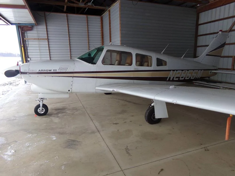 A side view of a white Piper Turbo Arrow III aircraft with brown and gold stripes, showing the registration N2855M, parked in a hangar.