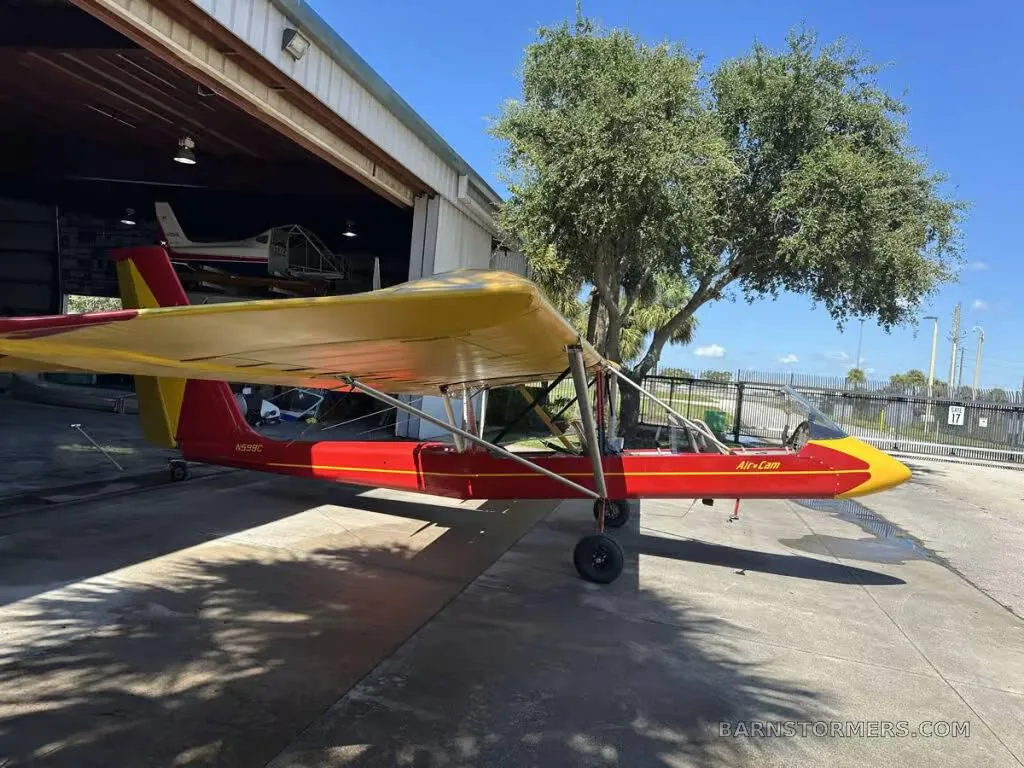 A red and yellow Air-Cam ultralight aircraft with registration N599C is parked on an airfield next to a tree.