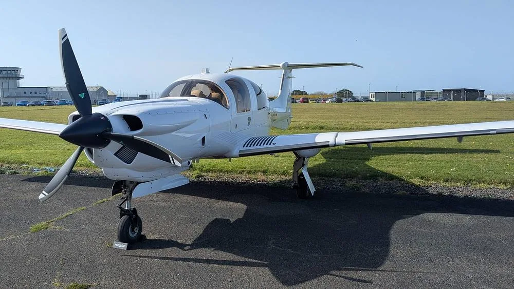 A white propeller aircraft with a black propeller on an airfield, viewed from the front-left.