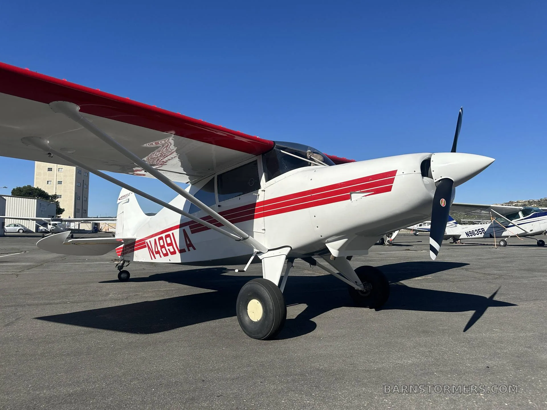 A white Maule M-7-235 aircraft with red stripes and registration N489LA is parked on an asphalt tarmac under a clear blue sky.